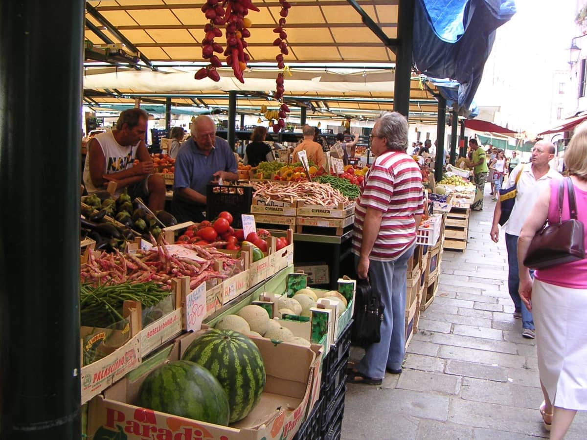 Frische Produkte und Gemüse auf dem Rialto-Markt in Venedig, mit Verkäufern und Käufern unter schattigen Markisen an einem belebten Morgen.