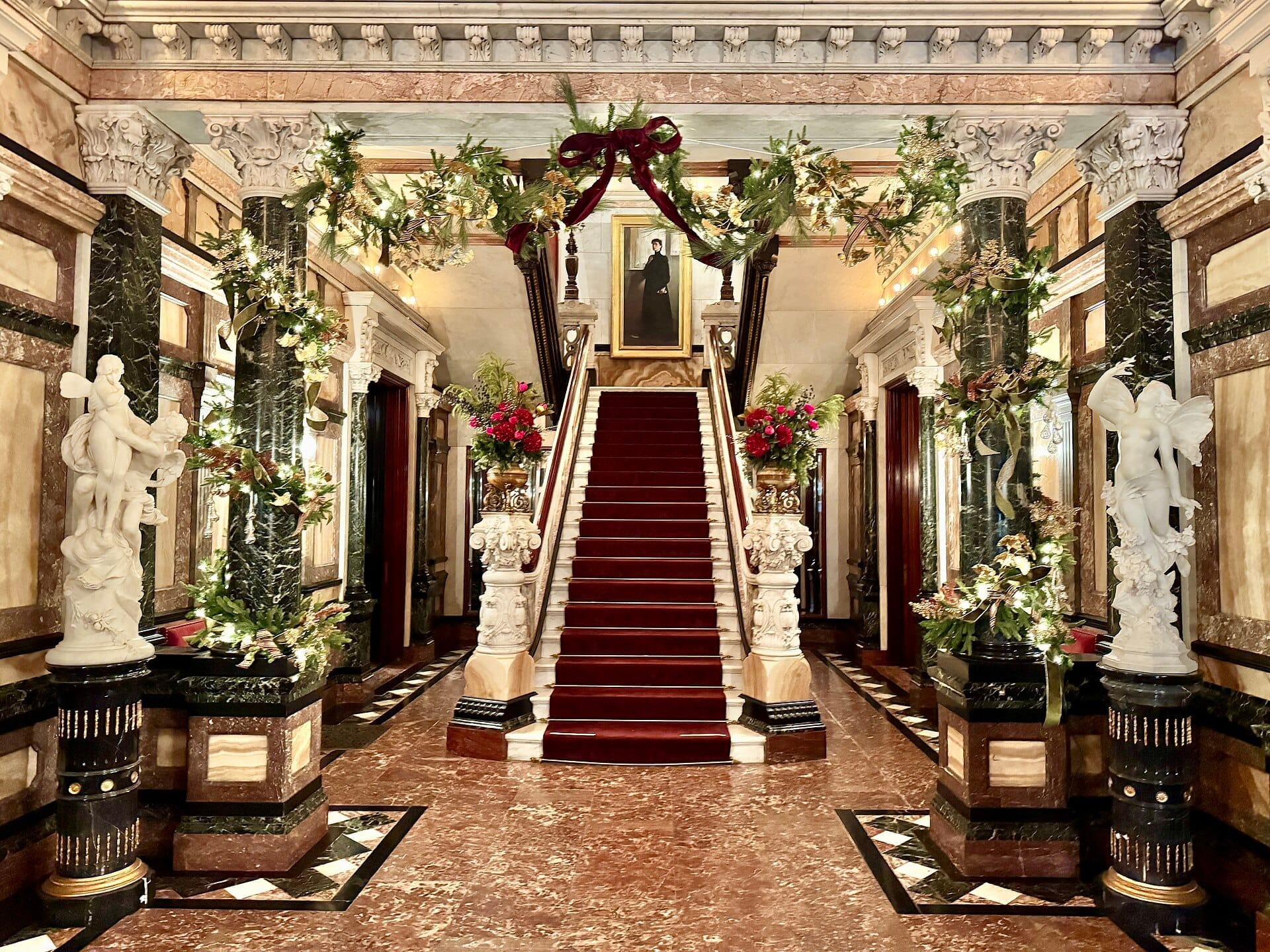 Grande ingresso del Richard H. Driehaus Museum con pavimenti in marmo, colonne ornate, decorazioni festive di piante e una scenografica scalinata con tappeto rosso.