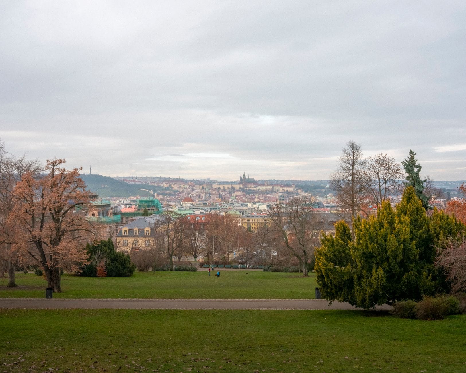 Weitläufige Parkwiese mit vereinzelten Bäumen im Vordergrund, mit Blick auf die Prager Stadtsilhouette und die entfernte Burg unter einem bewölkten Himmel.