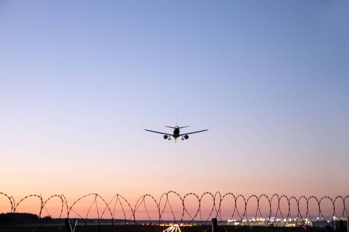 Ein Flugzeug im Landeanflug bei Sonnenuntergang, von hinten über einen Stacheldrahtzaun fotografiert, mit Flughafenlichtern und Terminal im Hintergrund.