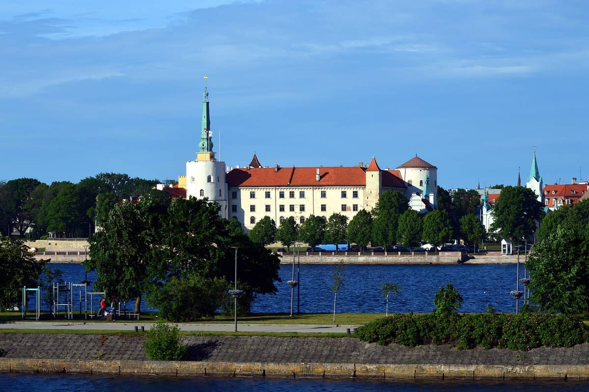 Das Rigaer Schloss mit seinen weißen Türmen, dem roten Dach und dem markanten Turm, fotografiert von der anderen Seite der Daugava an einem sonnigen Tag.