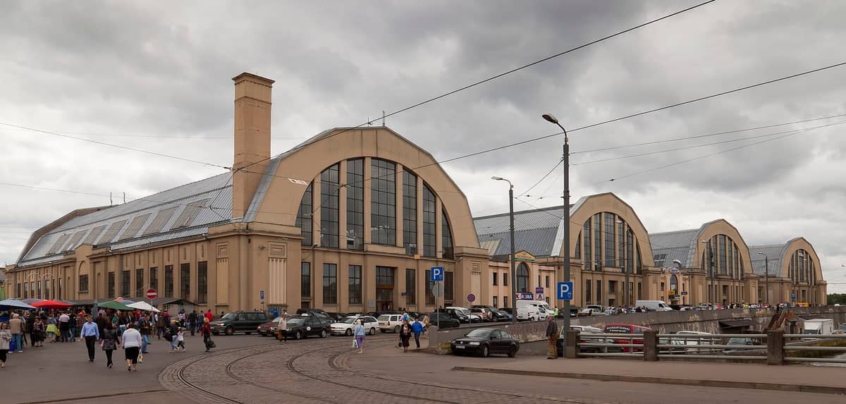 วิวภายนอกของอาคารโกดังโค้งอันเป็นเอกลักษณ์ของ Riga Central Market ในวันที่ท้องฟ้ามีเมฆ มีผู้คนและรถจอดอยู่ด้านหน้า