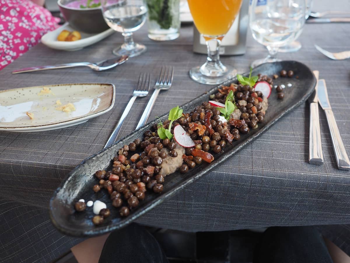 Nahaufnahme eines lettischen Restauranttisches in Riga mit einem langen Gericht aus grauen Erbsen und Speck, frischen Kräutern und Rettichgarnitur.
