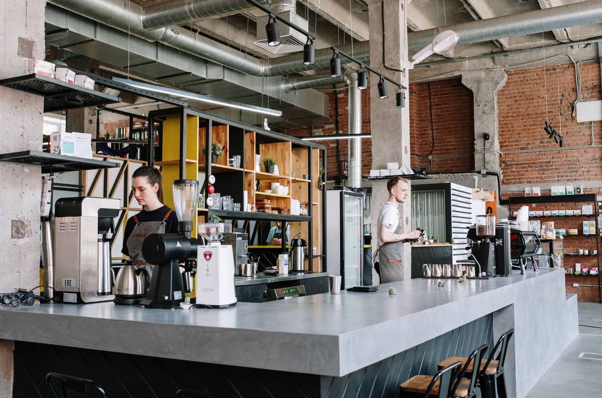 Modernes Café-Interieur in Riga mit Personal hinter einer geräumigen Theke, Kaffeeausrüstung, Regalen sowie Sichtmauerwerk und industriellem Dekor.