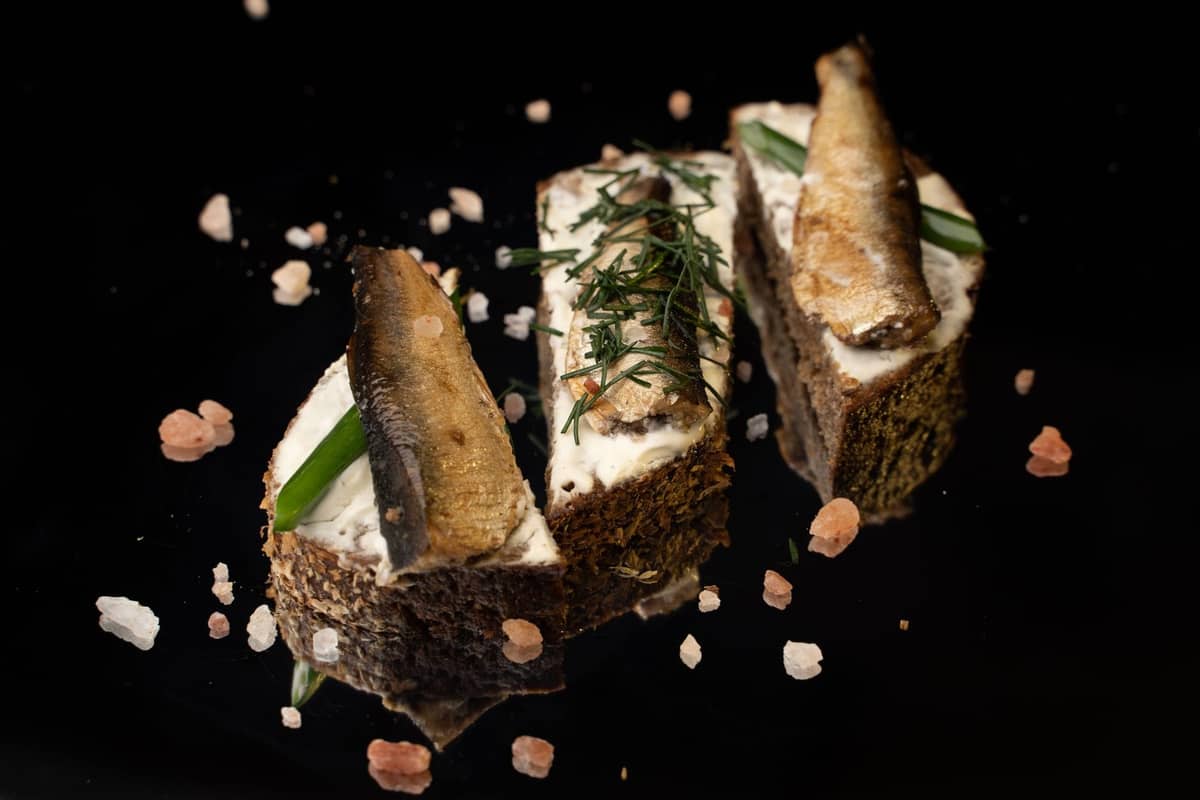 Scheiben dunkles Roggenbrot, belegt mit geräuchertem Fisch und frischen Kräutern auf schwarzem Hintergrund, mit gestreutem Salz.