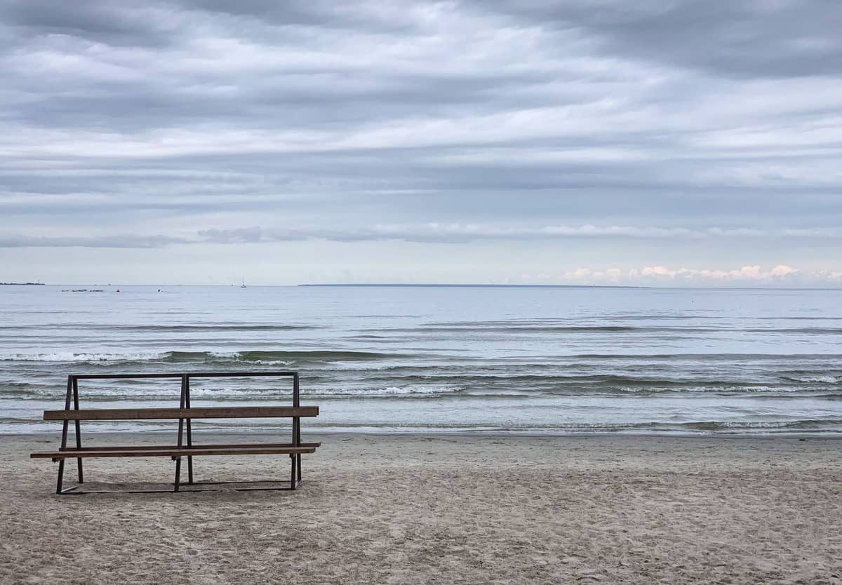 Leere Bank mit Blick auf einen ruhigen Sandstrand und die Ostsee unter einem bewölkten Himmel, die eine friedliche, romantische Atmosphäre heraufbeschwört.