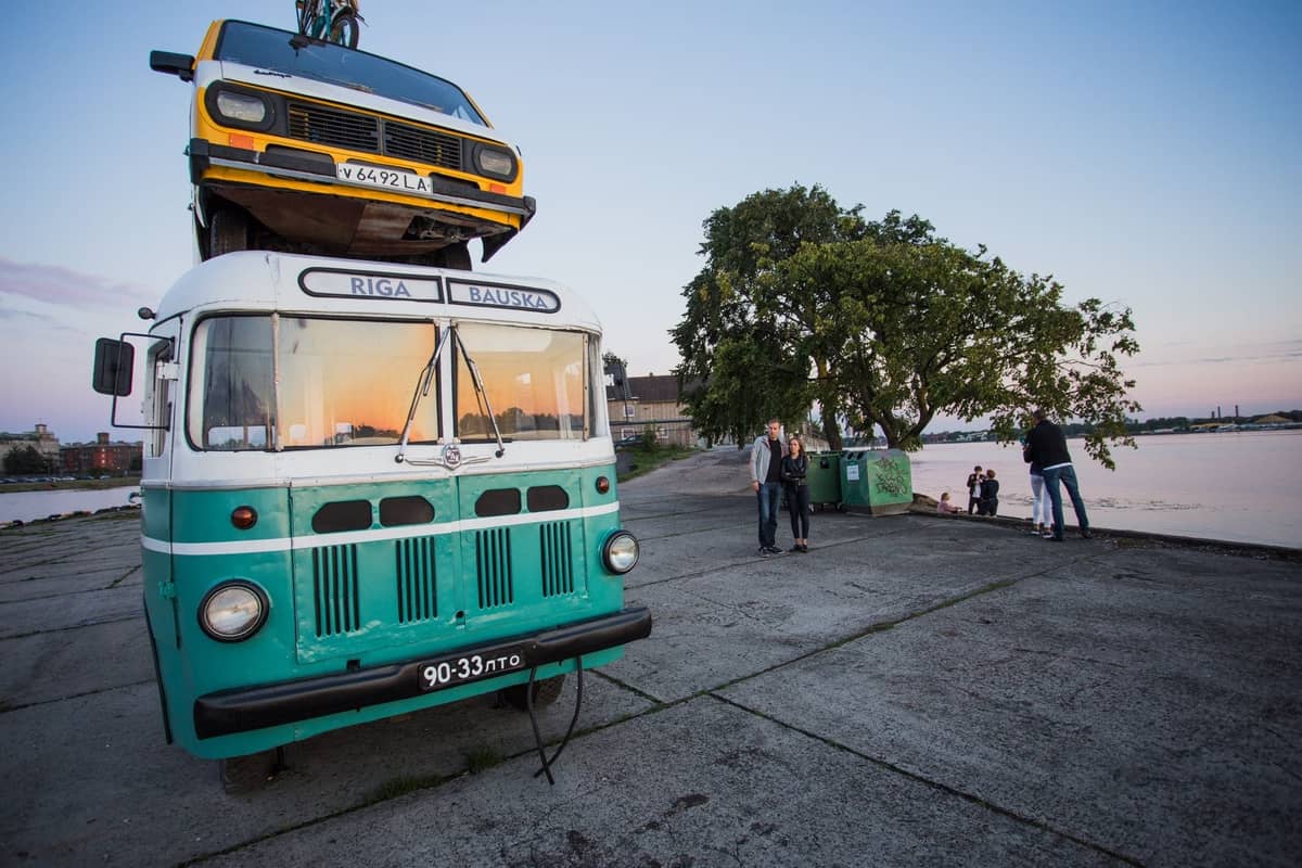 Ein alter grüner Riga-Bauska-Bus mit einem gelben Auto auf dem Dach, bei Sonnenuntergang am Ufer der Daugava geparkt, mit Paaren und Freunden in der Nähe.