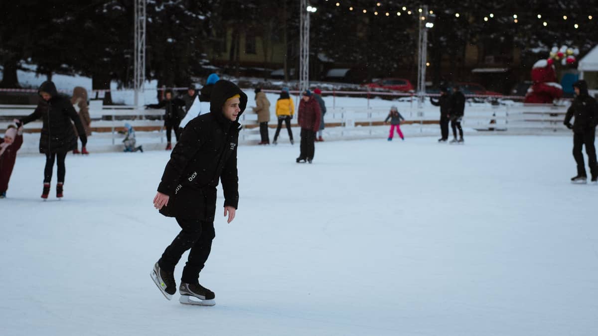 Menschen beim Schlittschuhlaufen im Freien im Winter auf einer beleuchteten Eisbahn, mit Bäumen und Stadtgebäuden im Hintergrund.