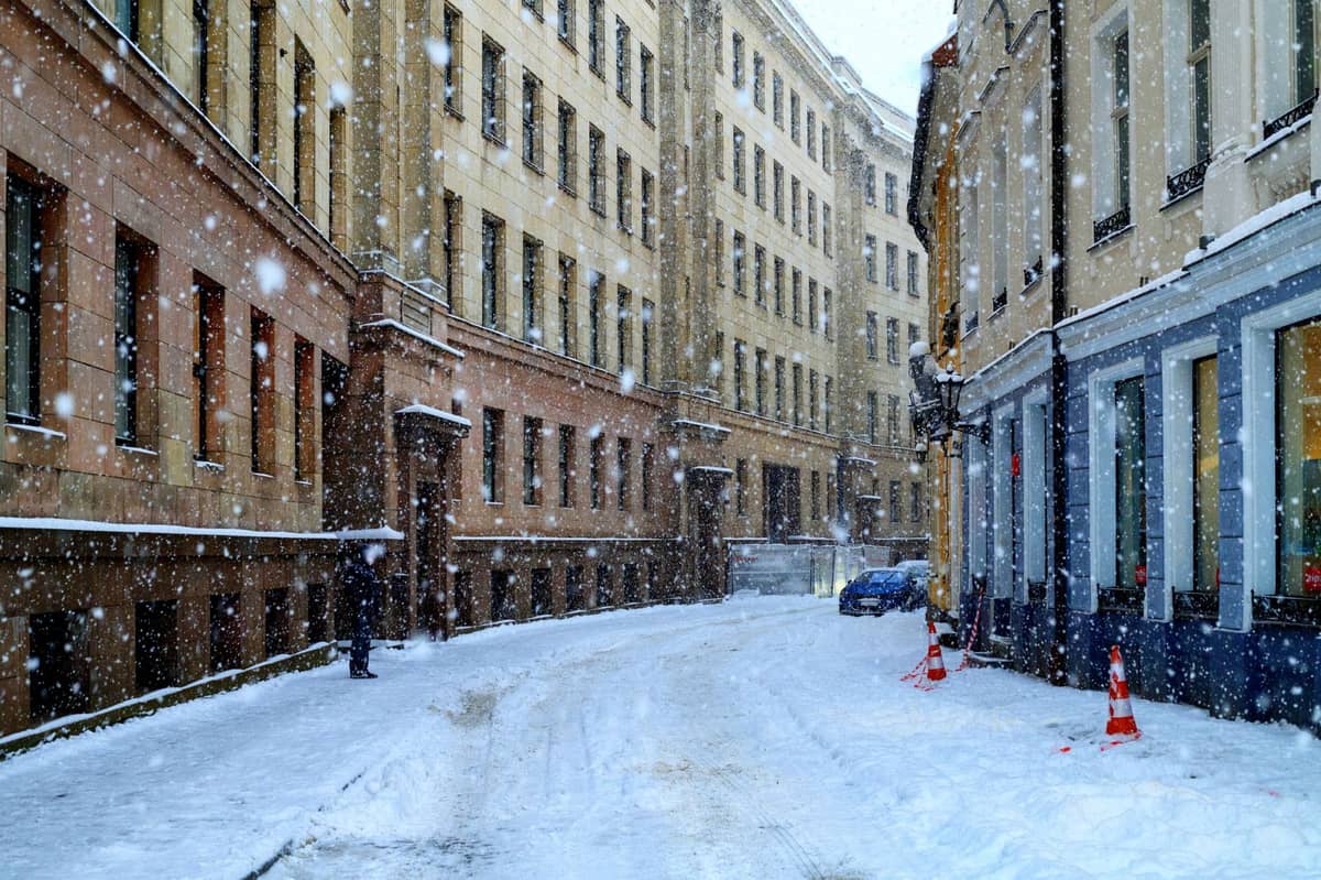 Schneefall auf einer ruhigen Straße, gesäumt von verzierten historischen Gebäuden im Winter, mit einigen Personen und Autos.