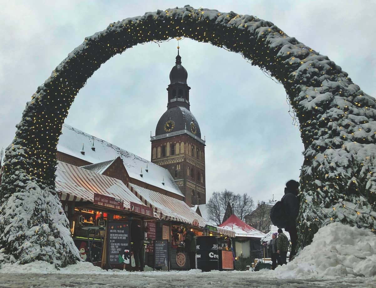 Ein verschneiter Eingangsbogen führt zum Rigaer Weihnachtsmarkt auf dem Domplatz in der Altstadt, mit festlich beleuchteten Ständen und dem Turm des Rigaer Doms im Hintergrund.