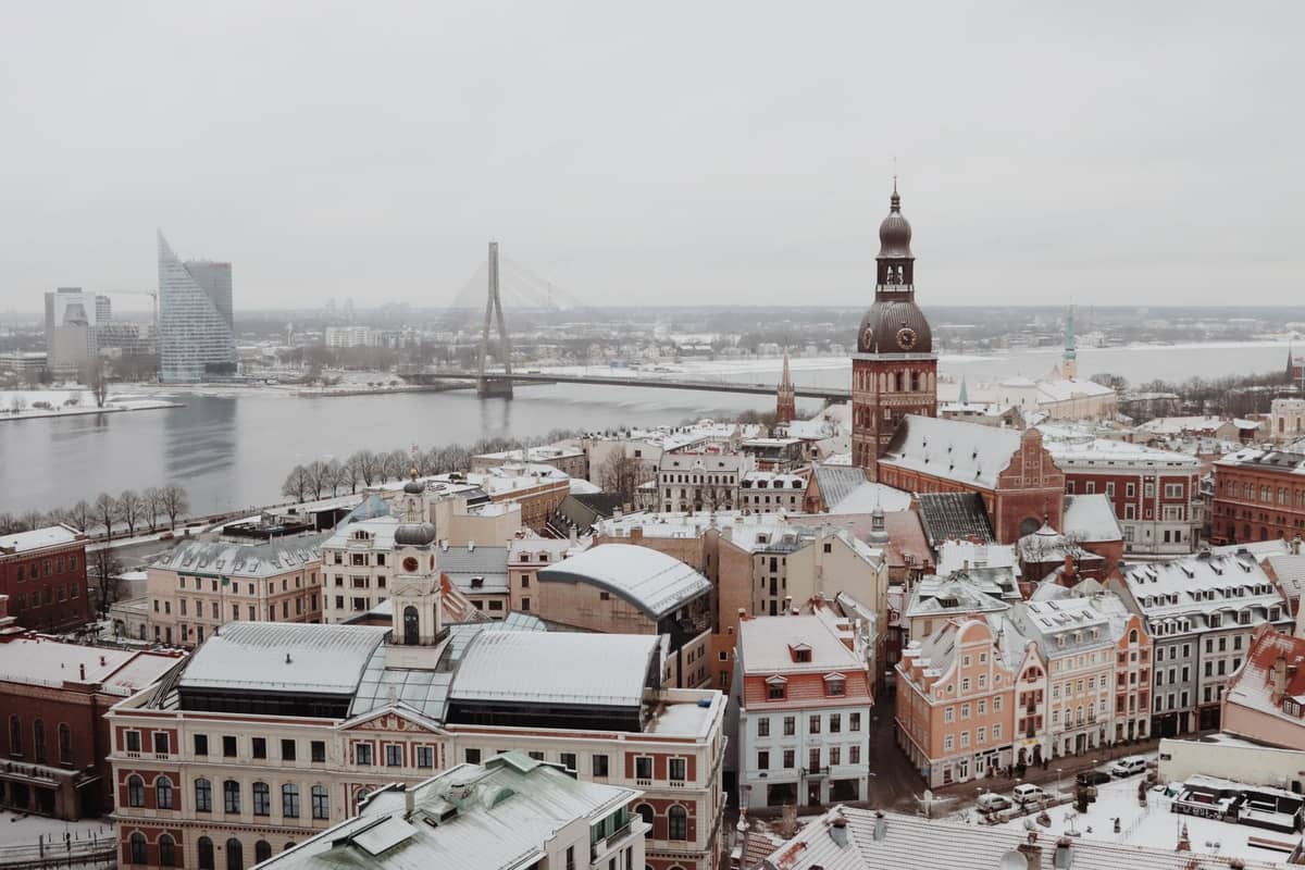 Ein weiter, bewölkter Winterblick auf Rigas Altstadt mit verschneiten Dächern, der Daugava und dem markanten Turm des Rigaer Doms.