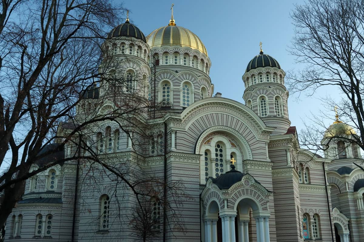 Weitwinkelansicht der neobyzantinischen Fassade und Kuppeln der Rigaer Christi-Geburt-Kathedrale mit goldenen Zwiebeltürmen, eingerahmt von kahlen Bäumen im Winterlicht.