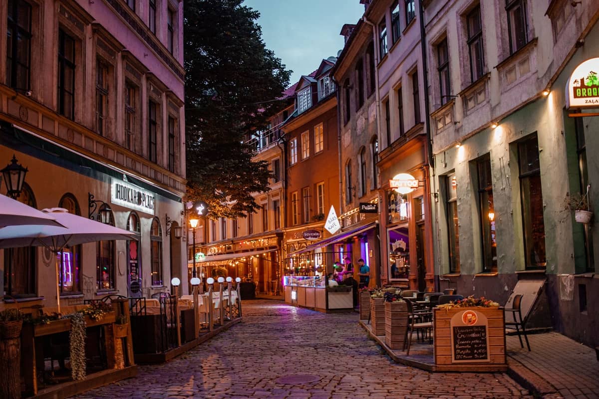 Abendliche Ansicht einer belebten Kopfsteinpflastergasse in Rigas Altstadt, gesäumt von Bars, Restaurants, Neonlichtern und Außensitzplätzen – Nachtleben-Atmosphäre pur.