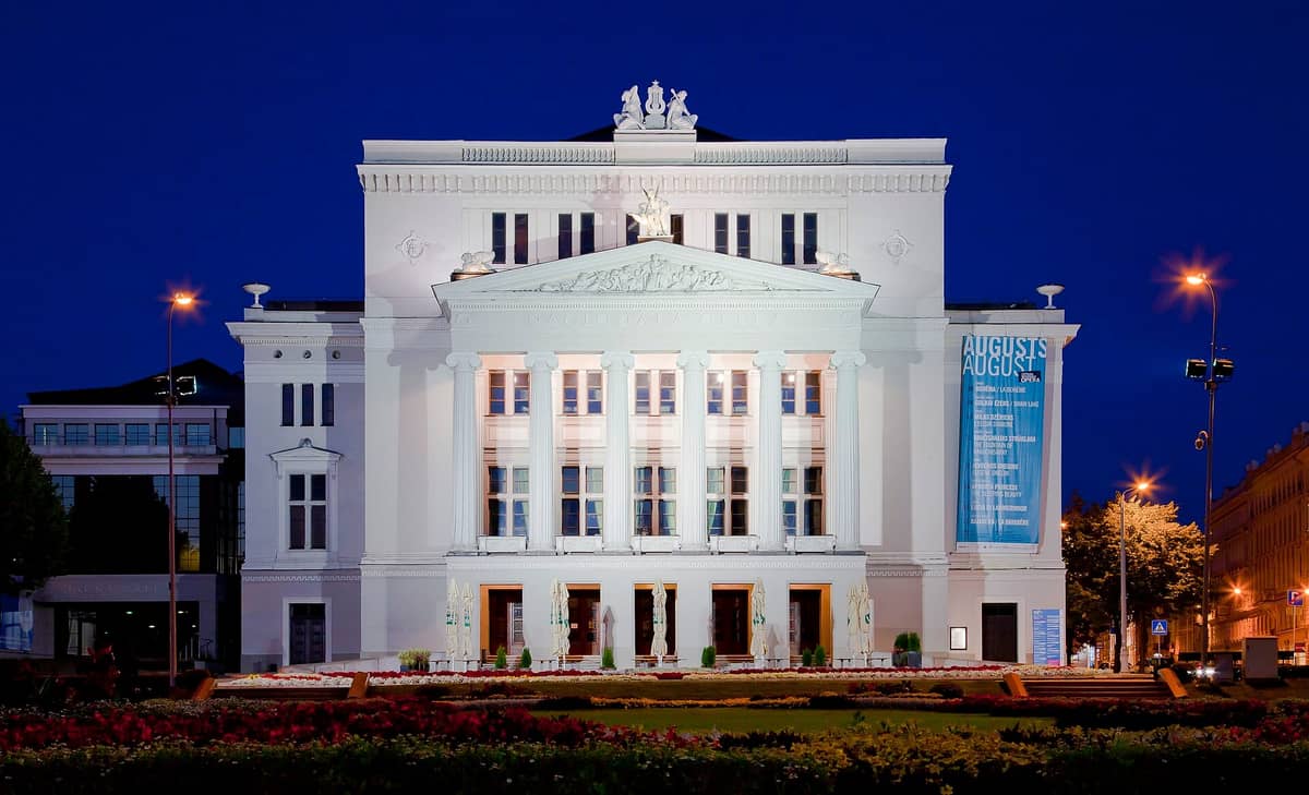 ด้านหน้าของ Latvian National Opera ที่สว่างไสวในยามค่ำคืนที่เมือง Riga พร้อมเสาคลาสสิกและรูปปั้นประดับตกแต่ง