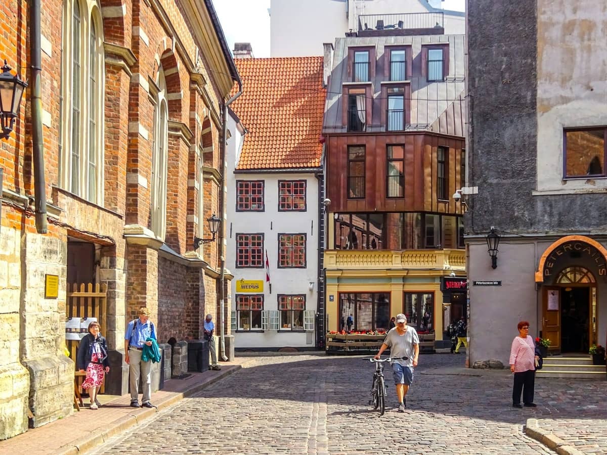 Kopfsteinpflasterstraße in der Altstadt von Riga mit historischen Backsteingebäuden, Passanten und moderner Architektur im Hintergrund bei hellem Sonnenschein.