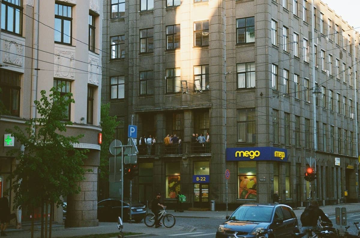 Straßenansicht im Zentrum von Riga mit Personen auf einem Apartmentbalkon über dem Supermarkt Mego, Radfahrern, Autos und klassischer Architektur im Abendlicht.
