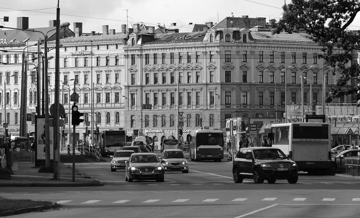 Belebte Kreuzung in der Stadt Riga mit Autos, Taxis, Bussen und historischen europäischen Gebäuden im Hintergrund bei Tageslicht.