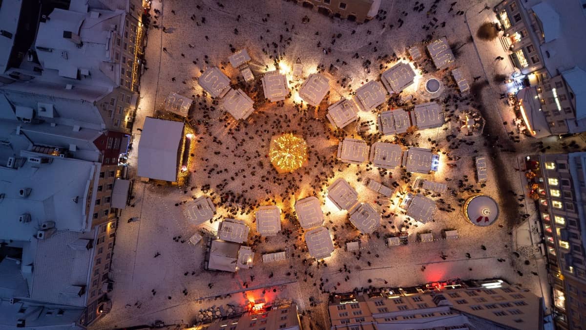 Luftaufnahme des Altstadtplatzes von Riga im Winter mit Weihnachtsmarktständen, festlichen Lichtern und Menschenmassen rund um einen dekorierten zentralen Bereich.