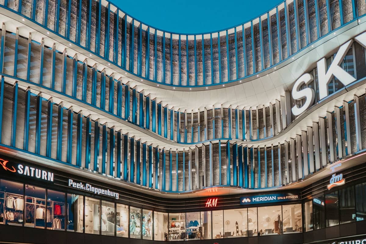 Zeitgenössische Einkaufszentrumfassade in Riga mit beleuchteten Geschäften wie H&M, Saturn und Peek & Cloppenburg am Abend.
