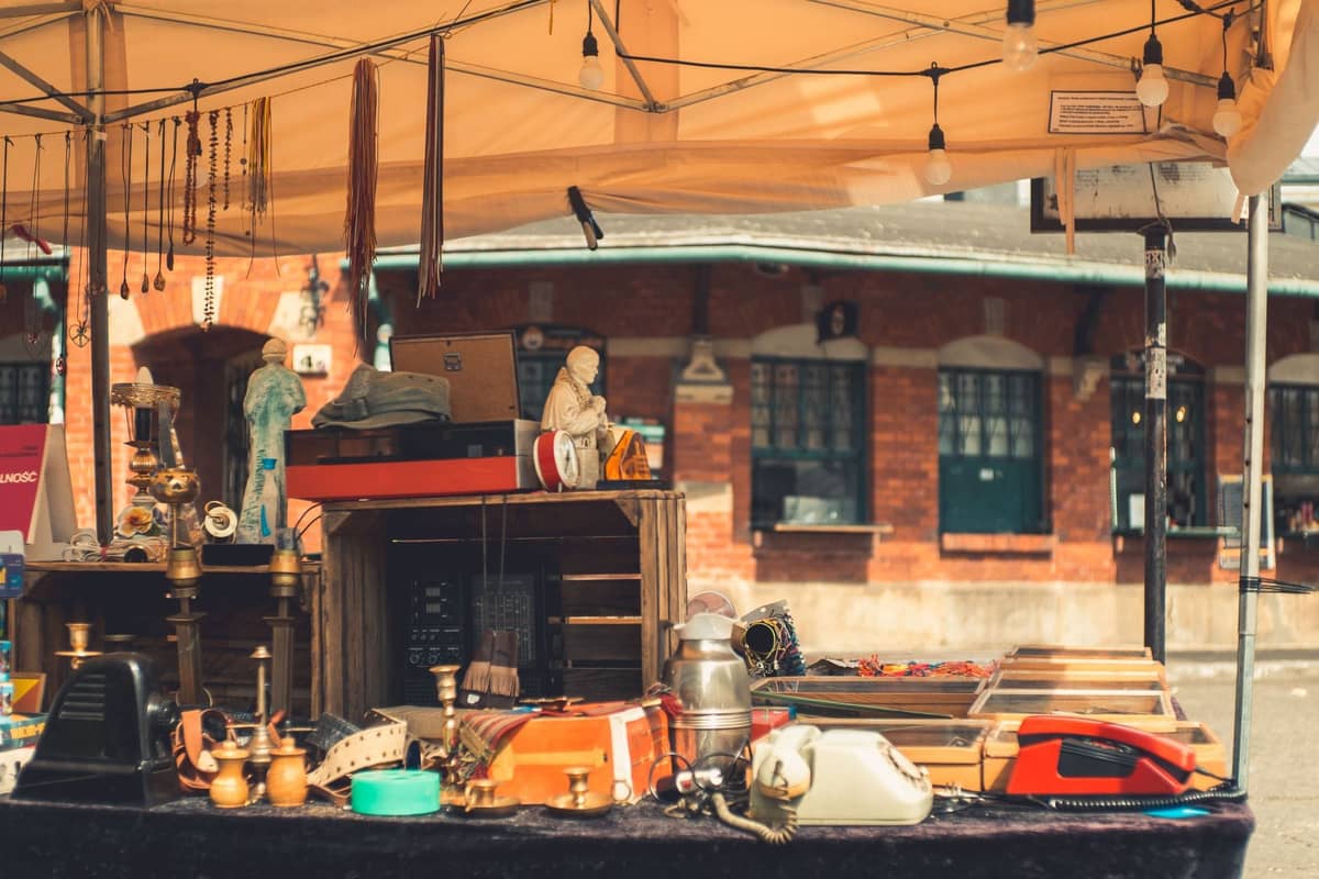 Flohmarktstand im Freien vor einem Backsteingebäude, der Vintage-Artikel wie Telefone, Geschirr und Figuren unter einem Sonnendach an einem sonnigen Tag ausstellt.