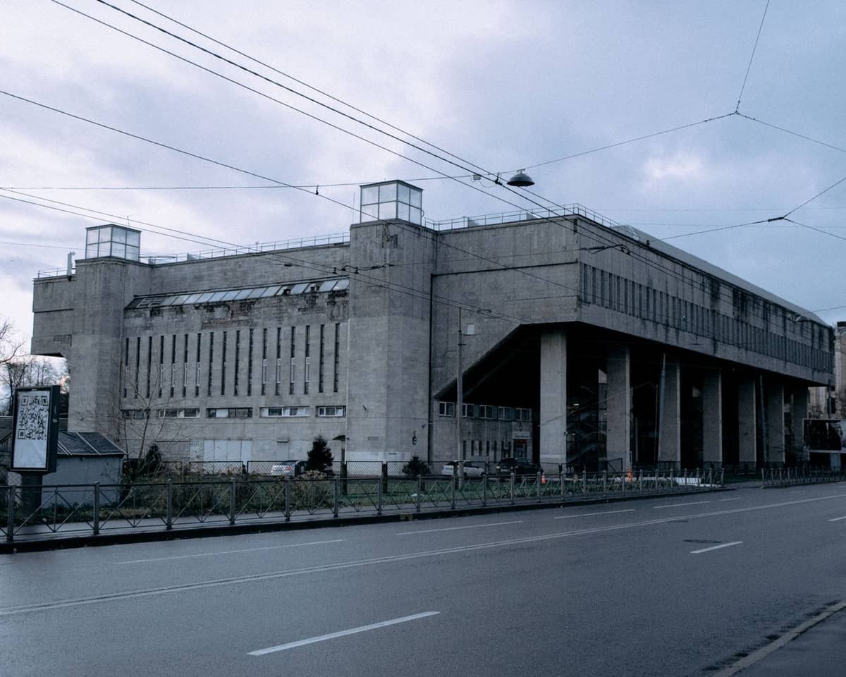 Großes Betongebäude im sowjetischen Brutalismus-Stil in Riga, vermutlich das ehemalige Druckhaus der Lettischen Akademie der Wissenschaften, mit Straße und Straßenbahnleitungen im Vordergrund bei bewölktem Himmel.