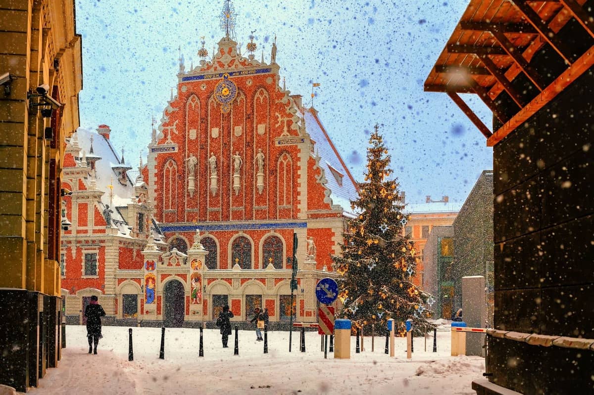 Schwarzhäupterhaus am Rathausplatz in Riga während eines Schneefalls, mit festlichen Weihnachtslichtern an einem Baum und Menschen, die über den verschneiten Platz gehen.