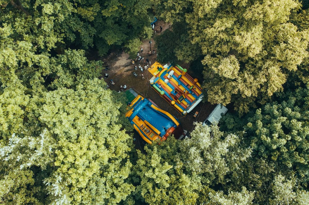 Luftaufnahme eines bewaldeten Parkgeländes mit bunten aufblasbaren Spielgeräten zwischen den Bäumen und spielenden Kindern im Freien