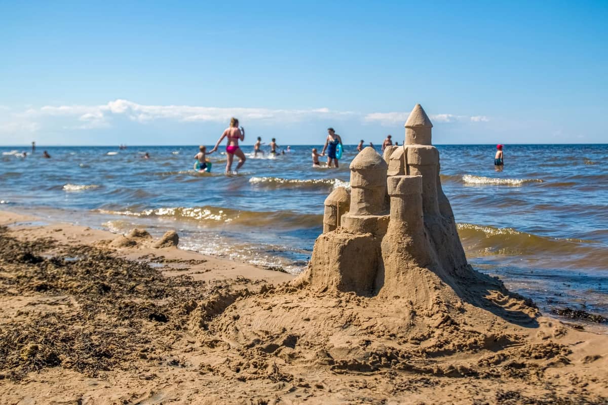 Familien und Kinder, die im Meer spielen und schwimmen, in der Nähe einer großen Sandburg an einem breiten Sandstrand unter blauem Himmel.
