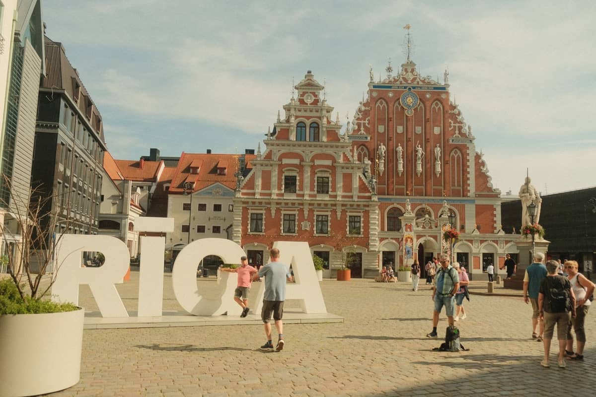 Große weiße RIGA-Buchstaben vor dem verzierten Schwarzhäupterhaus in Rigas Altstadt, mit Menschen, die einen sonnigen Tag genießen.