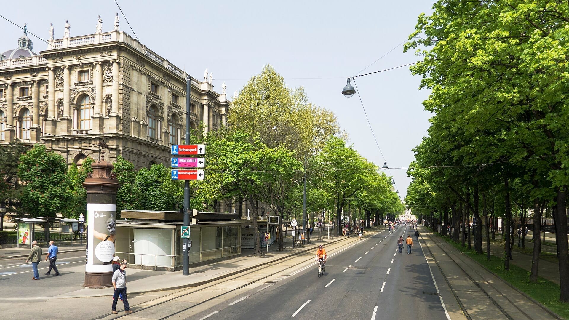 Weitwinkelansicht der Wiener Ringstraße mit einem historischen Steingebäude, baumsgesäumtem Boulevard, Wegweisern, Radfahrern und Fußgängern an einem klaren Tag.
