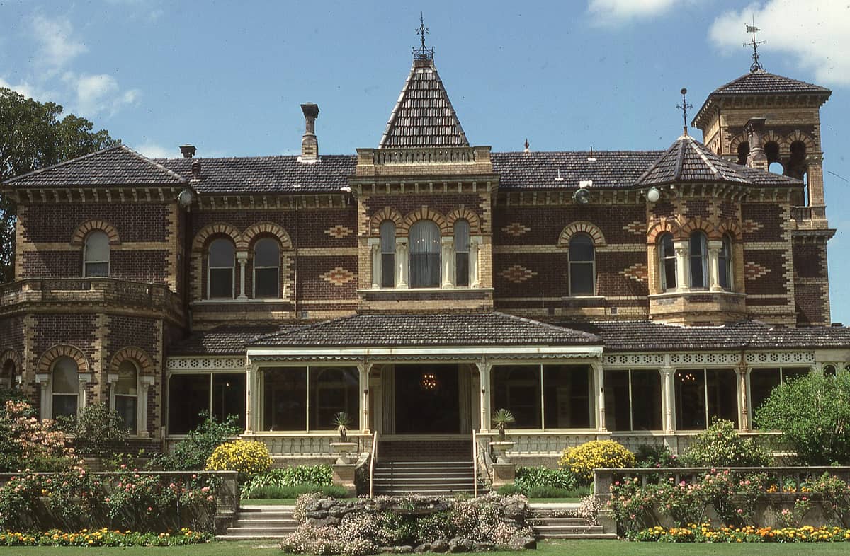 Weitwinkelperspektive auf das Rippon Lea Estate – ein imposantes viktorianisches Herrenhaus in Melbourne mit aufwendigem Ziegelmauerwerk, Veranda, Türmen und üppigen Gärten unter blauem Himmel.