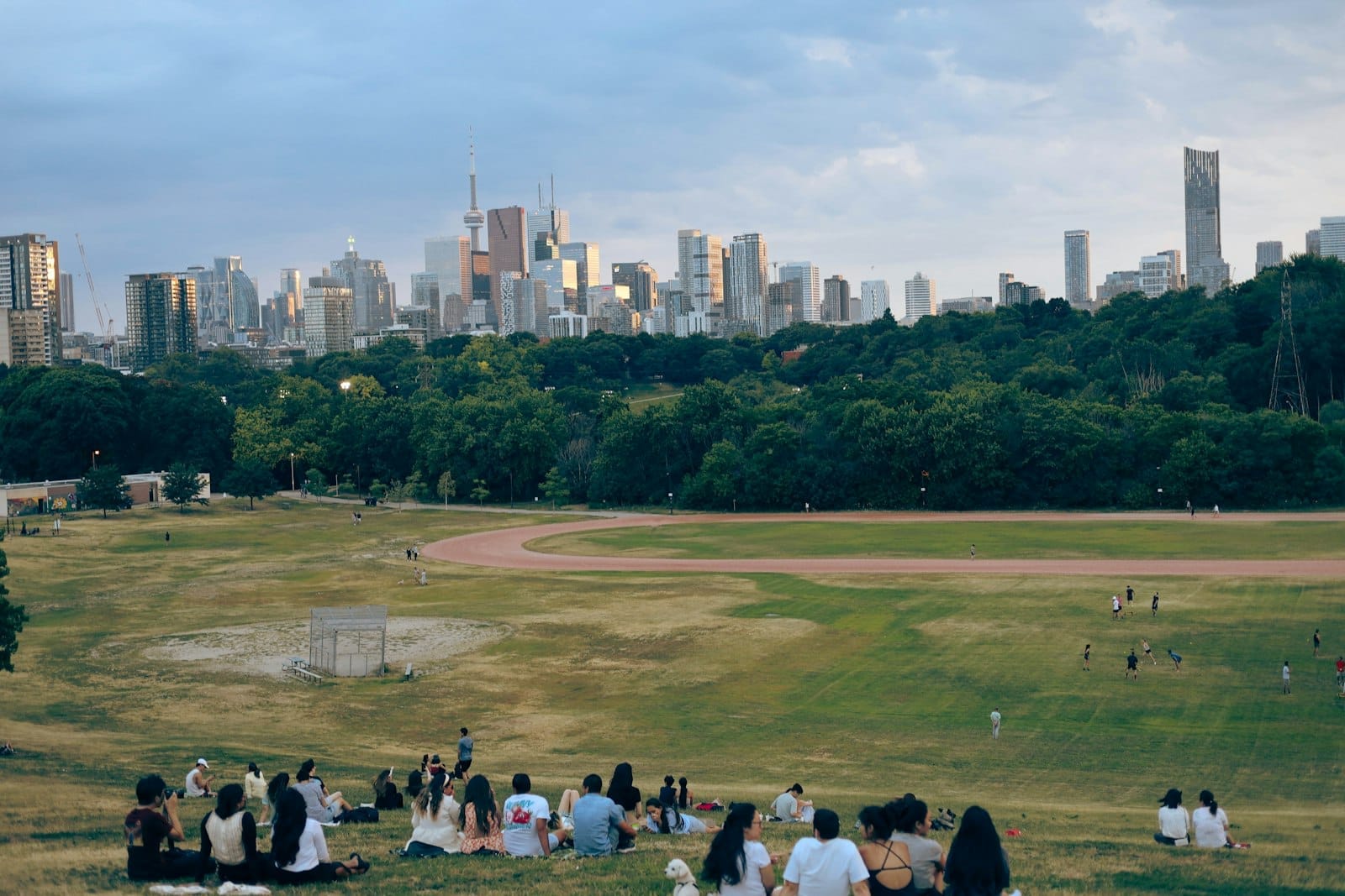 วิวของ Riverdale Park East ในโตรอนโต มีผู้คนนั่งพักผ่อนบนเนินหญ้า พร้อมเส้นขอบฟ้าของใจกลางเมืองเป็นฉากหลังในยามพลบค่ำ