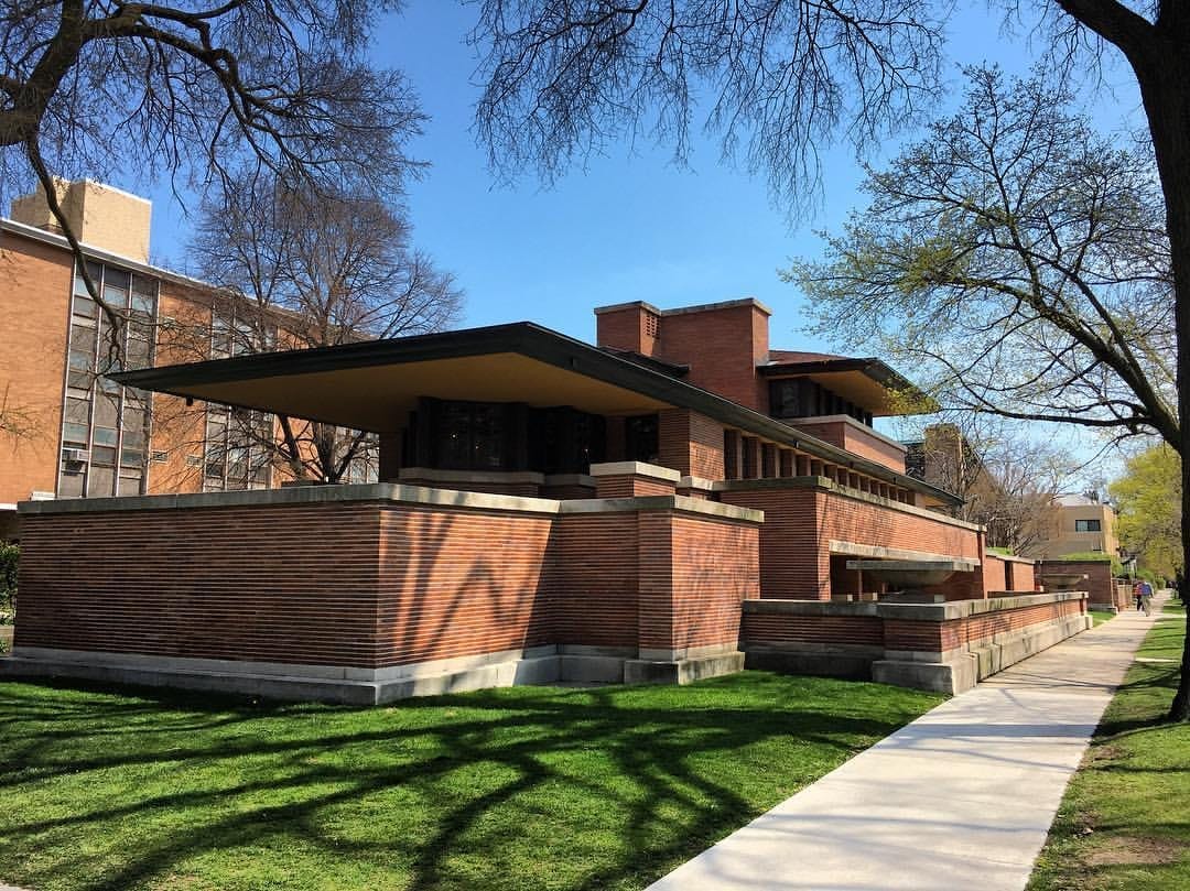 Robie House a Hyde Park, un imponente edificio in mattoni basso con linee orizzontali, circondato da prato verde e alberi primaverili sotto un cielo azzurro limpido.