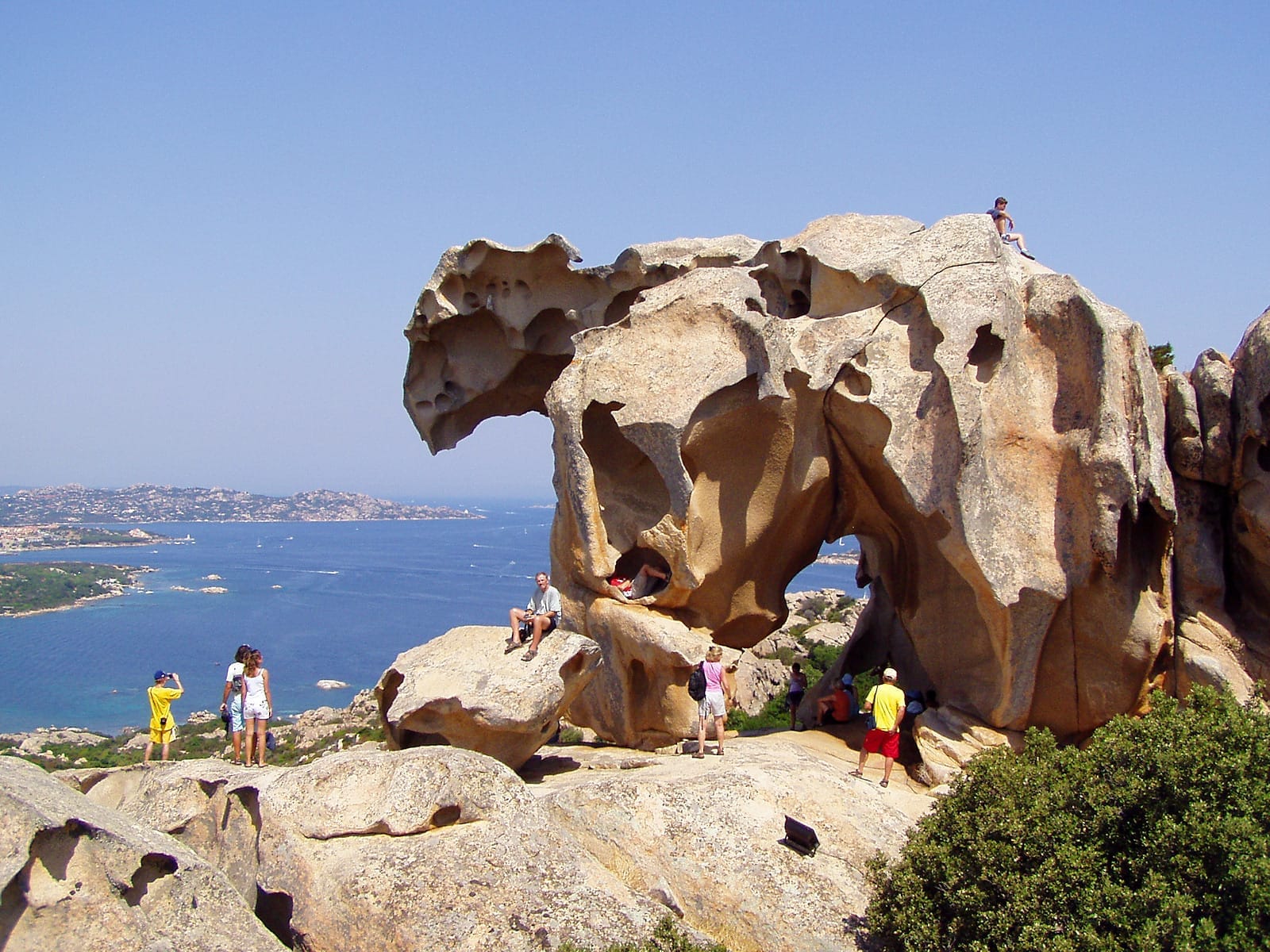 Touristen erkunden die Roccia dell'Orso, eine vom Wind geformte Granitfelsformation mit Blick auf die blaue Küste und die üppigen grünen Hügel von Palau, Sardinien.
