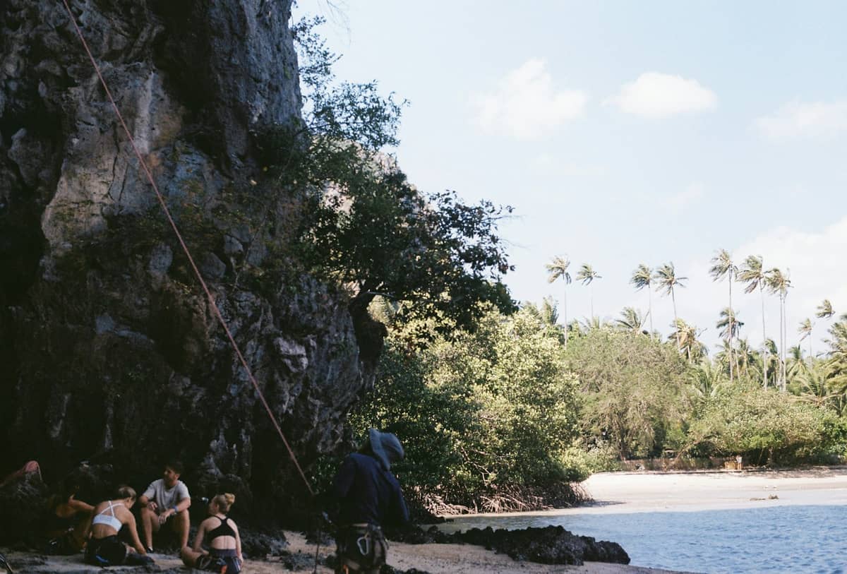 Grupa wspinaczy odpoczywających przy wapiennych klifach z linami asekuracyjnymi na Railay Beach w Krabi, otoczonych tropikalnymi drzewami i błękitnym niebem.