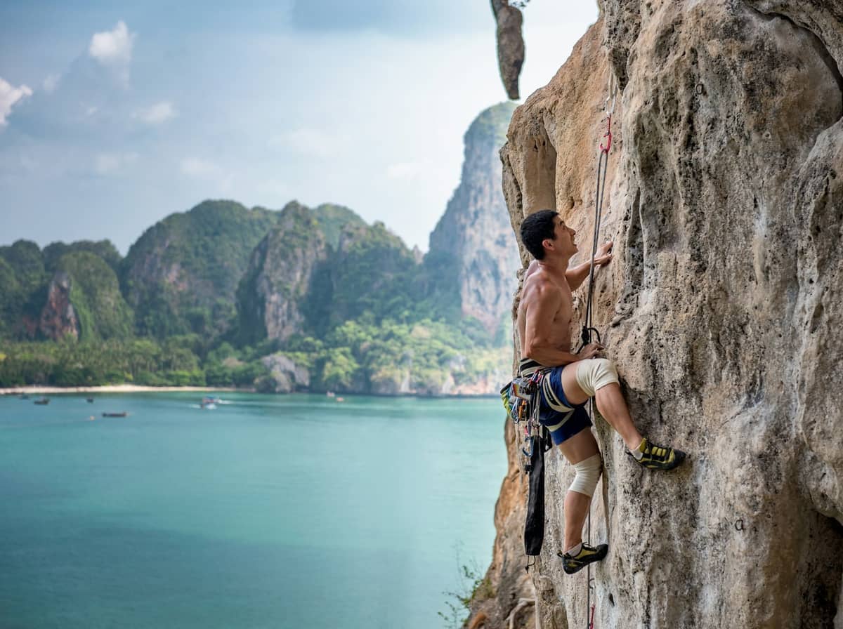 Mężczyzna wspinający się na skałę wapienną na Railay Beach, Krabi, na tle turkusowego morza i imponujących zielonych gór.