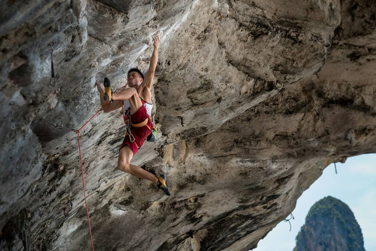 Wspinacz w czerwonych szortach wspina się na strome wapienne klify w Railay, Krabi, z linami i tropikalną scenerią w tle.