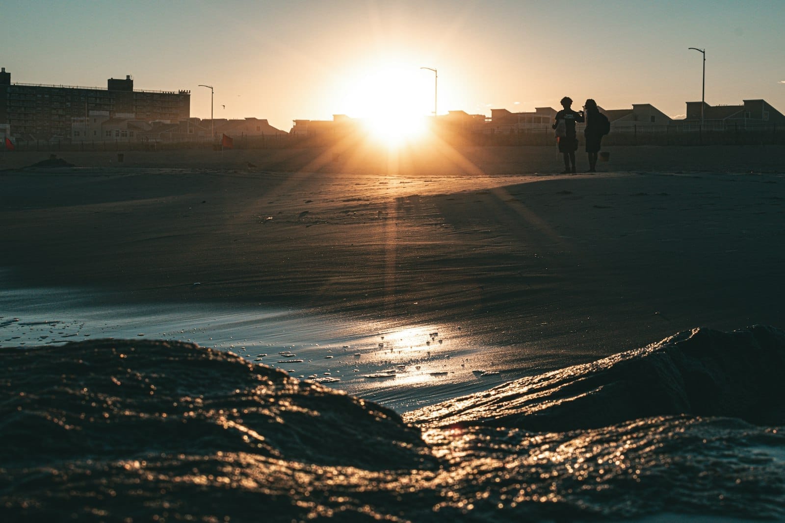 Due persone passeggiano sulla spiaggia sabbiosa al tramonto, con bassi edifici sagomati contro il sole dorato e l'Oceano Atlantico che scintilla in primo piano.
