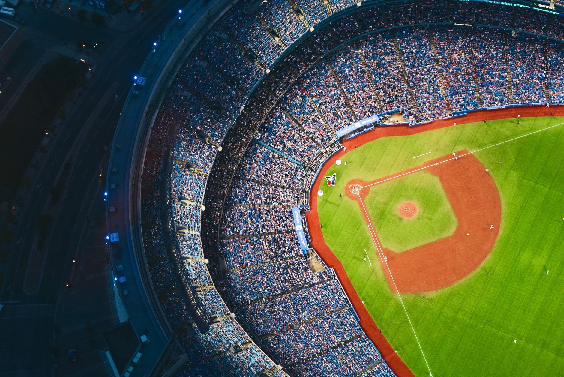 มุมมองทางอากาศของ Rogers Centre ที่เต็มไปด้วยแฟนๆ โดดเด่นด้วยสนามเบสบอลสีเขียวสดใส ที่นั่งในสนามกีฬาสีสันสดใส และถนนโดยรอบของโตรอนโตในยามค่ำคืน