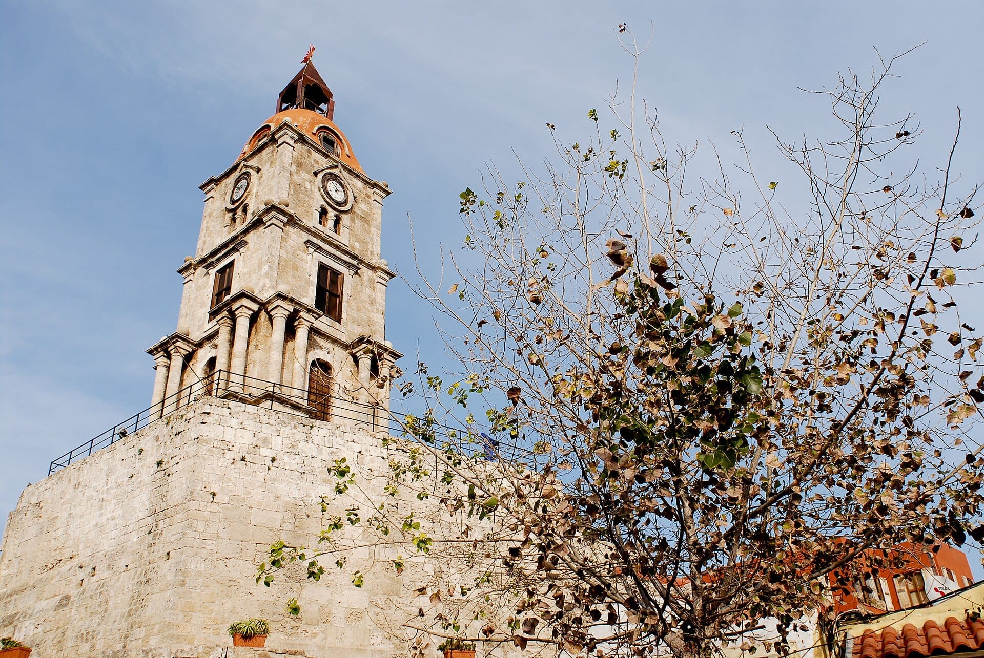 หอนาฬิกา Roloi ในเมืองเก่า Rhodes ตั้งตระหง่านเหนือกำแพงหินข้างต้นไม้ใบหนาทึบ ใต้ท้องฟ้าสีฟ้าใส