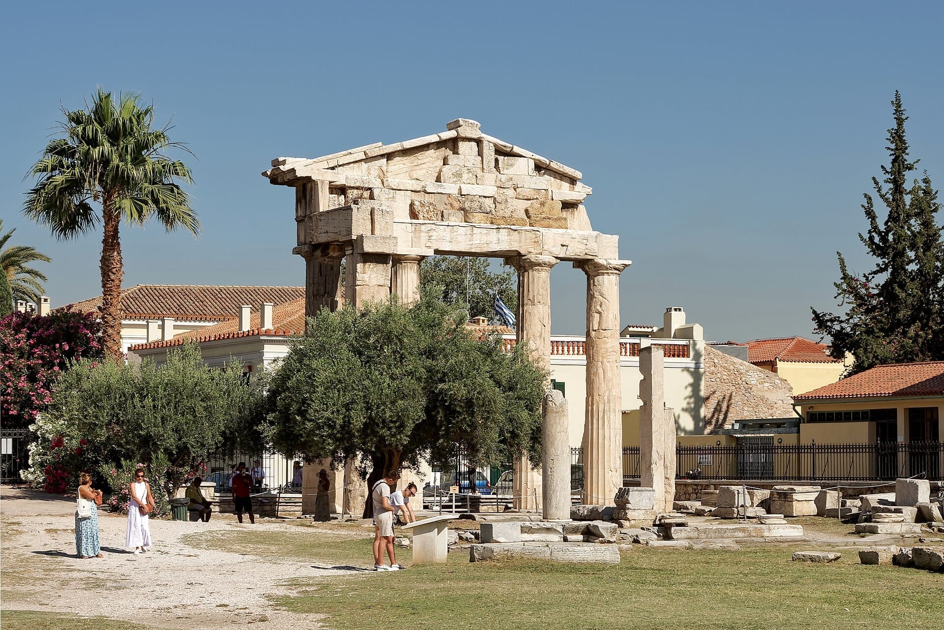 La Porta di Atena Archegetis nell'Agorà Romana di Atene, circondata da ulivi, palme e visitatori sotto un cielo azzurro limpido.