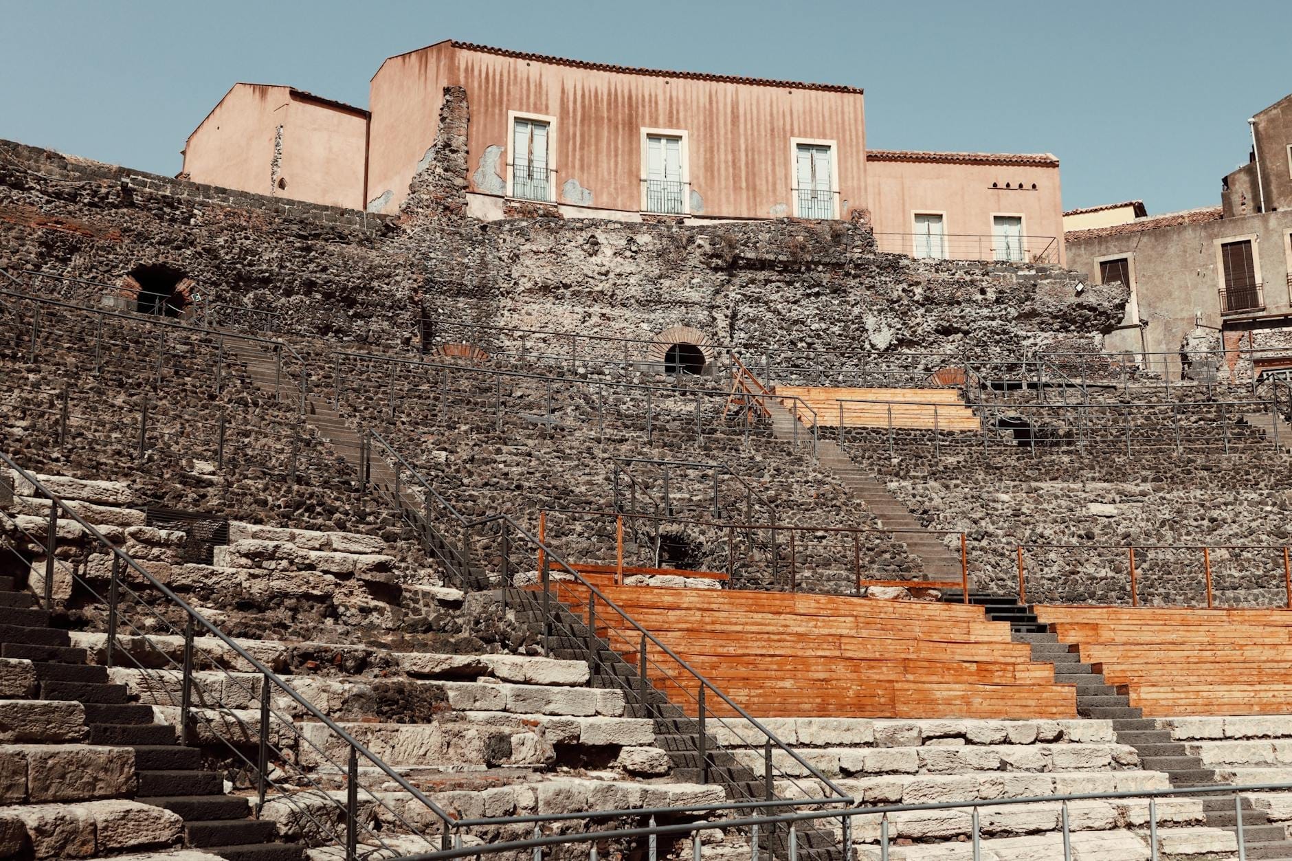 Die steinernen Sitzreihen des Römischen Amphitheaters von Catania ragen unter den Gebäuden der Innenstadt hervor – antike Ruinen im Kontrast zur modernen Stadt, unter klarem Himmel.