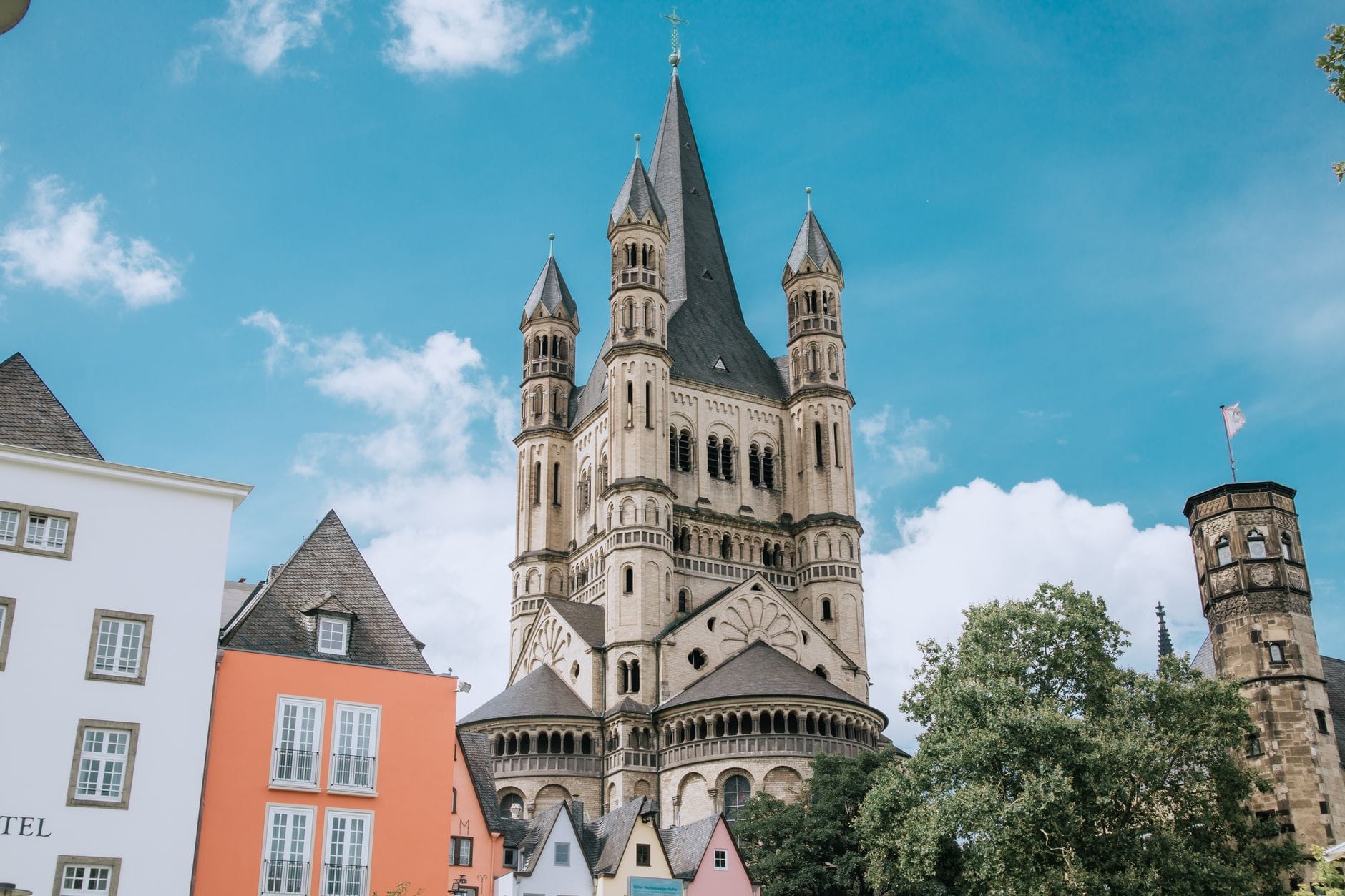โบสถ์โรมาเนสก์ขนาดใหญ่ในเมือง Cologne ที่มีหลายหอคอยและหน้าต่างโค้ง ตั้งอยู่เบื้องหลังอาคารเมืองเก่าสีสันสดใสใต้ท้องฟ้าสีฟ้า