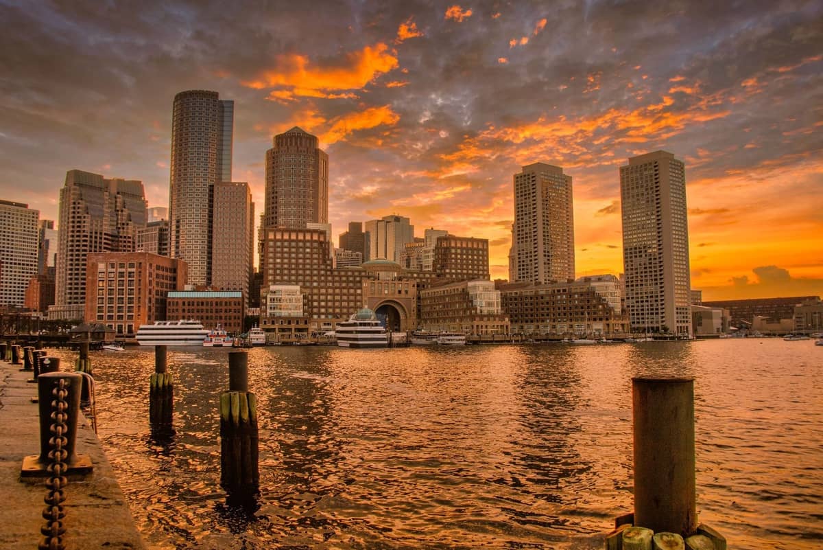Boston Harbor bei Sonnenuntergang mit Stadtsilhouette, leuchtendem Himmel sowie Booten und Kreuzfahrtschiffen am Ufer.