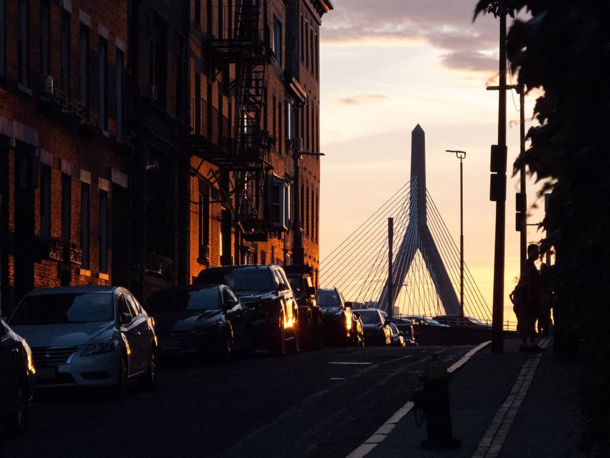 Straße in Boston bei Sonnenuntergang mit der Zakim Bridge im Hintergrund und Fußgängern, die eine romantische Stadtatmosphäre heraufbeschwören.