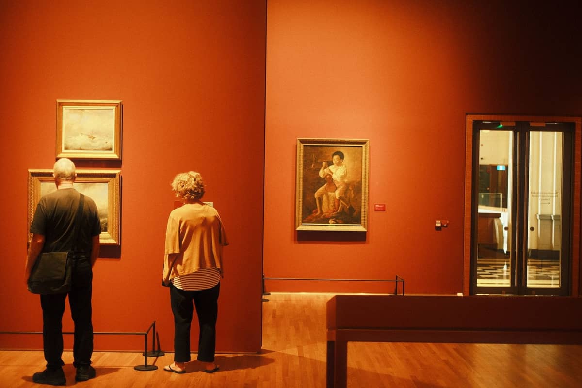 Ein Paar steht in einem warmbeleuchteten Kunstmuseumsaal und bewundert gerahmte Gemälde an tiefroten Wänden.