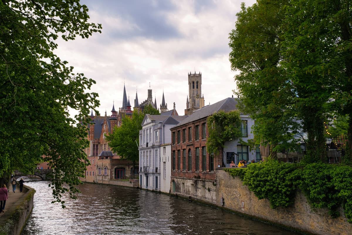 Canal in Bruges lined with medieval buildings, trees, and the Belfry tower visible in the background on a cloudy day.