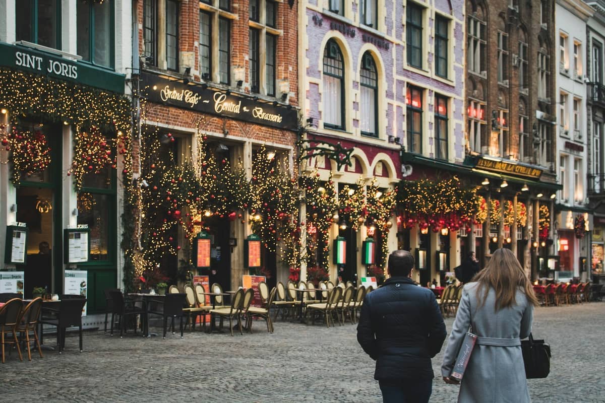 Colorful Bruges street with festive holiday lights, decorated cafes like Sint Joris and Grand Café Central, empty outdoor tables, and two people walking by.