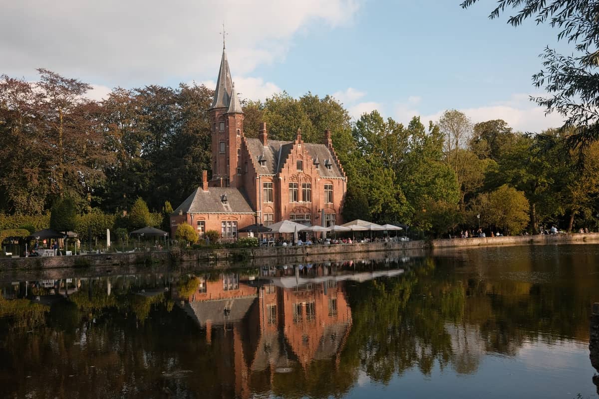 A romantic, fairy-tale style brick manor house beside Minnewater lake in Bruges, reflecting on calm water with trees in background.