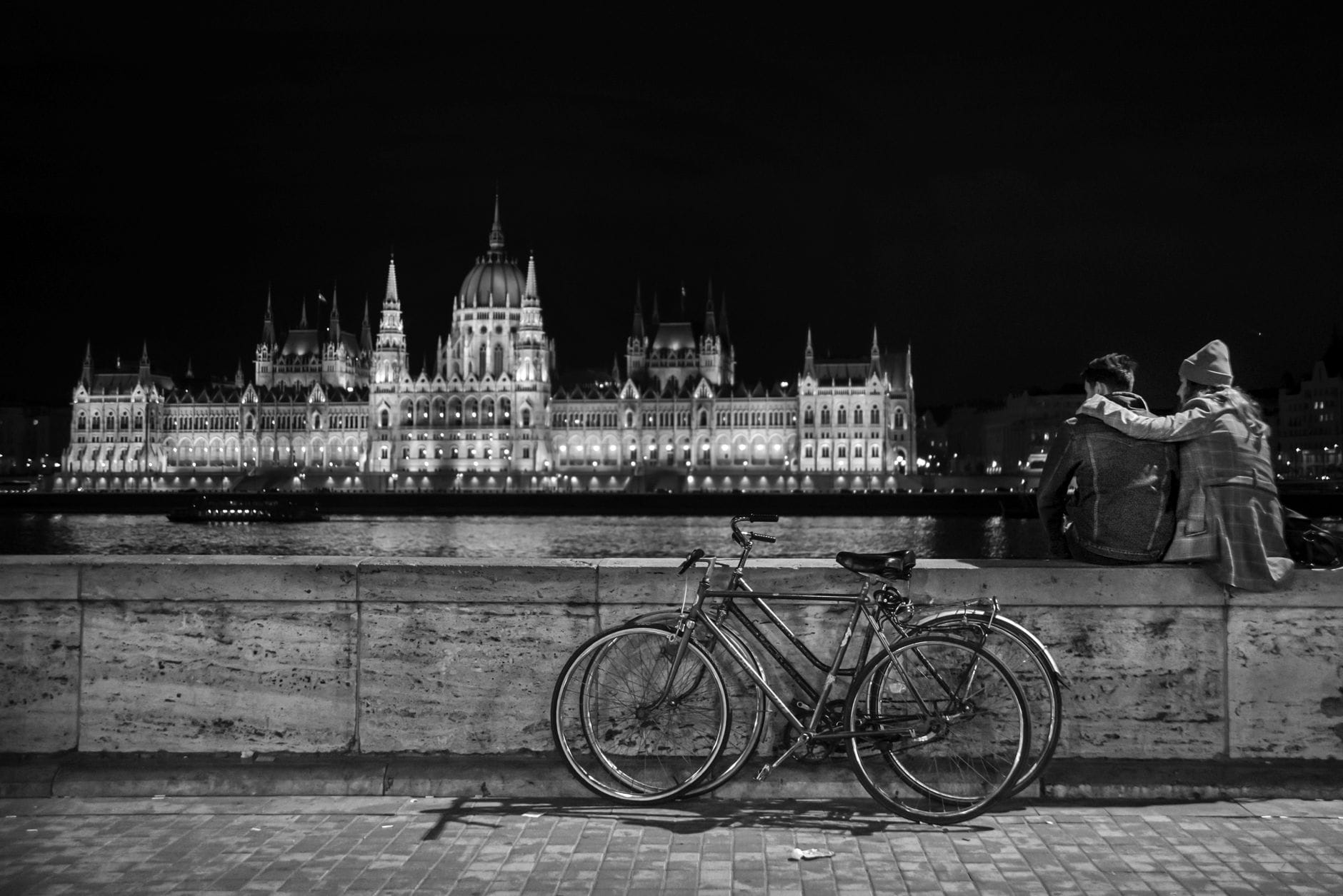 Pärchen mit Fahrrädern am Flussufer bei Nacht, das Ungarische Parlamentsgebäude prachtvoll beleuchtet auf der anderen Seite der Donau – eine romantische und ikonische Stadtansicht.